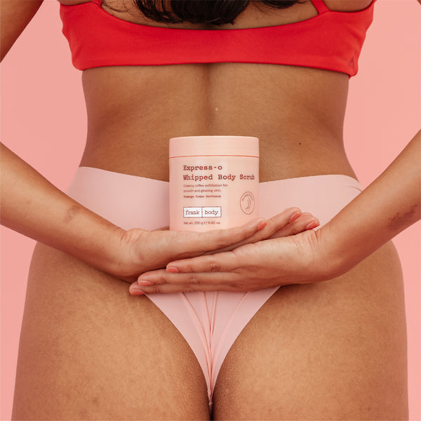 A model in a coral red bikini top and light pink underwear holds a jar of Express-o Whipped Body Scrub against their lower torso, set against a solid pink background.