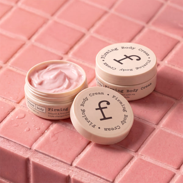 Three jars of Firming Body Cream arranged on pink square tiles, with one open jar showing the pale pink cream inside and two closed jars showing the cream-colored lids with black text.