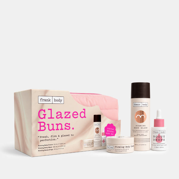 Frank Body Glazed Buns set featuring a pink and beige gift box, a large smoothing body butter bottle, a smaller body butter jar, a serum bottle, and a dropper bottle, arranged on a light gray background.