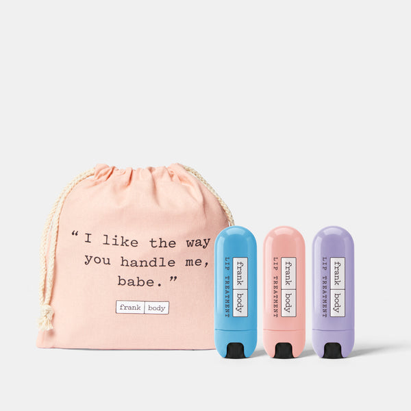 Three lip balm tubes in blue, pink, and purple are displayed next to a light pink drawstring bag with a quote printed on it, all on a light gray background.