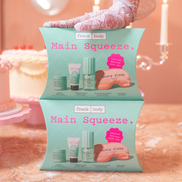 Two stacked Frank Body Main Squeeze gift boxes held by a gloved hand, featuring illustrated skincare products and a pink bootie sleep mask on a peach-toned background with candles and a blurred cake.