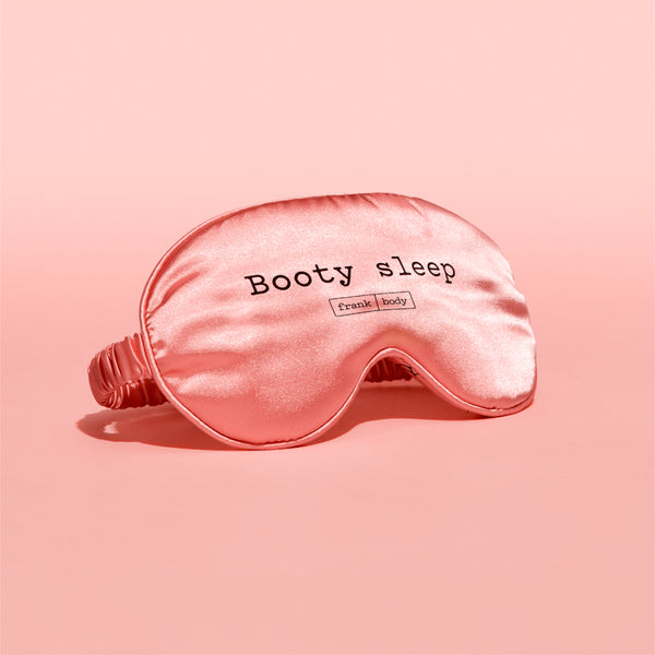 A pink satin sleep mask with the words 'Booty sleep' and a small 'Frank Body' logo printed on the front, set against a solid pink background.