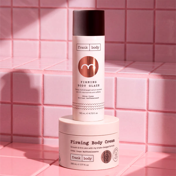 A Frank Body Firming Body Glaze spray canister and a matching Firming Body Cream jar, both in soft pink packaging with dark brown caps, displayed on a pink tiled surface.