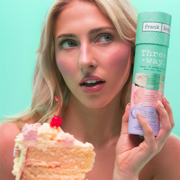 A blonde woman holding a slice of pink cake and a mint green aerosol can labeled 'frank body Threeway' against a solid mint green background.