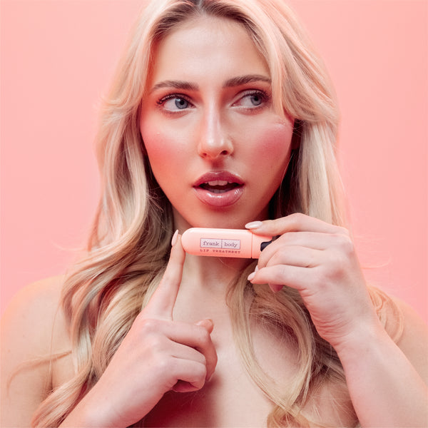 A blonde woman holding a peach-colored lip product tube near her face against a solid pink background.