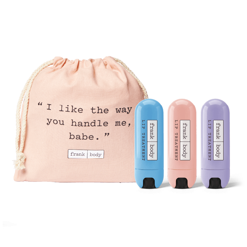 Three lip treatment stick tubes in blue, pink, and purple are displayed in front of a blush pink drawstring pouch with a humorous printed quote. The tubes are labeled 'Frank body' and 'lip treatment'.