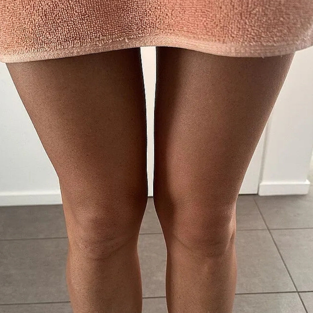 Close-up of a person's legs wearing sheer, skin-toned hosiery or tights, with a peach-colored textured fabric at the top, standing indoors on a gray tiled floor.