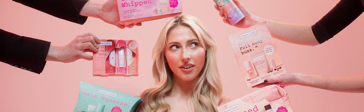 Blonde model surrounded by hands holding up frank body holiday gift sets against a pink background.