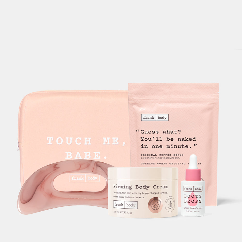 A Frank Body self-care set featuring a blush pink pouch with 'TOUCH ME, BABE.' text, a matching pink curved tray, a cream jar labeled 'Firming Body Cream,' a small white bottle labeled 'BOOTY DROPS,' and a pink stand-up pouch with a quote, arranged on a light gray background.